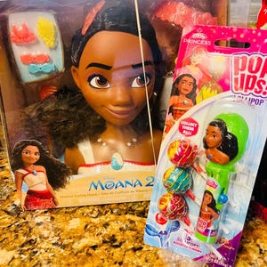 May include: A Disney Moana styling head doll in a box, with accessories. Next to it is a package of Pop Ups! lollipops featuring Moana. The lollipop package has assorted flavors and a Moana character on the handle.