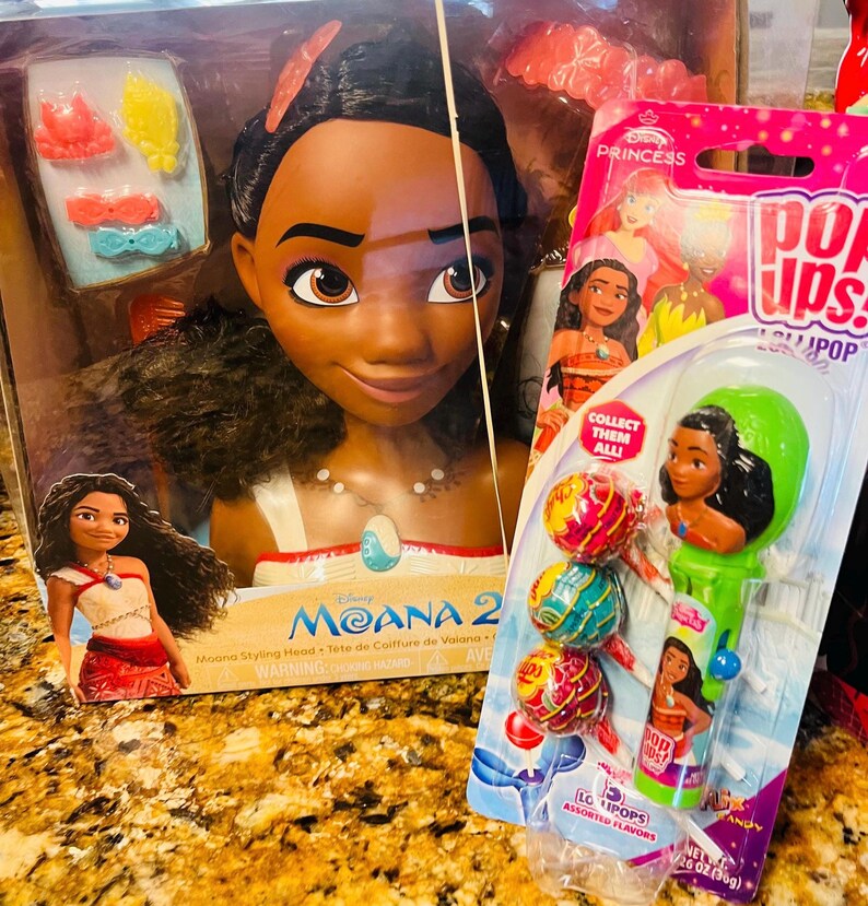 May include: A Disney Moana styling head doll in a box, with accessories. Next to it is a package of Pop Ups! lollipops featuring Moana. The lollipop package has assorted flavors and a Moana character on the handle.