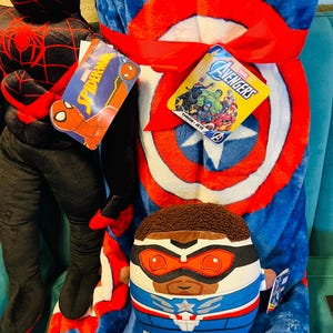 May include: A blue Captain America throw blanket with a red and white shield design is tied with a red ribbon. A plush Falcon character and a Spider-Man plush are also visible. Tags with Marvel branding are attached.