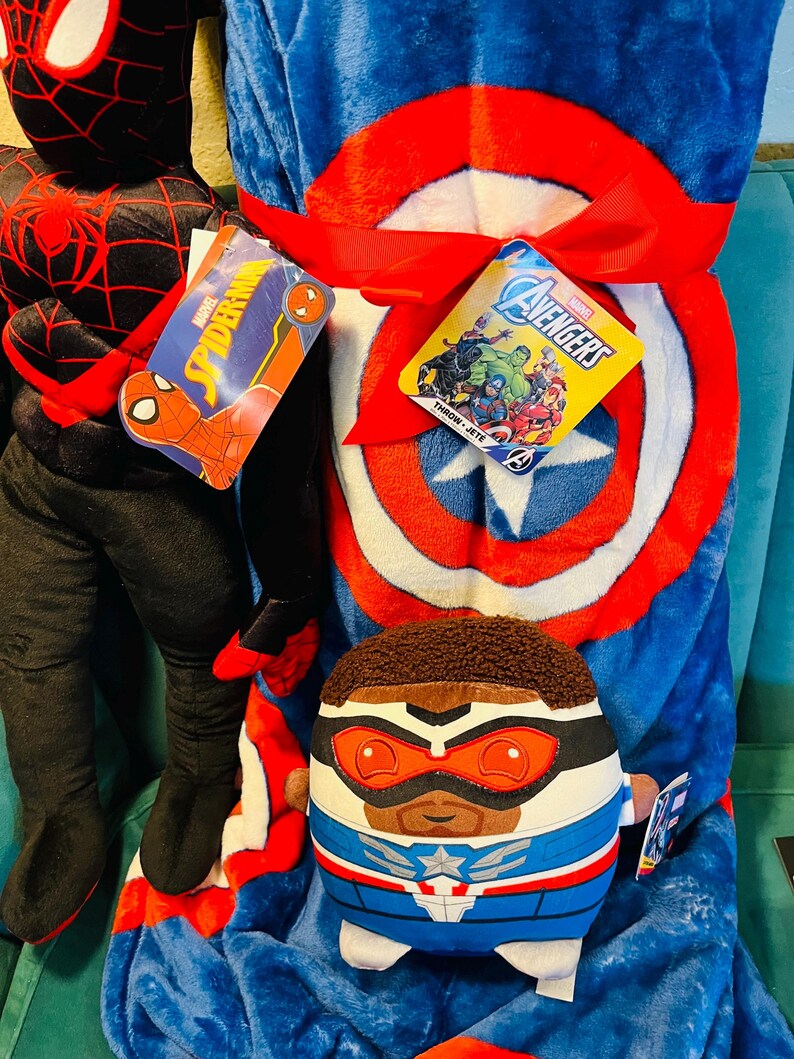 May include: A blue Captain America throw blanket with a red and white shield design is tied with a red ribbon. A plush Falcon character and a Spider-Man plush are also visible. Tags with Marvel branding are attached.