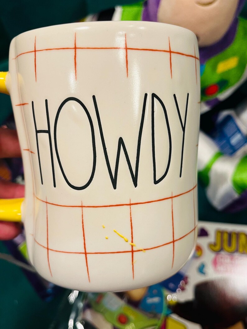 May include: White ceramic mug with the word "HOWDY" in black lettering. The mug has a yellow handle and a red grid pattern. There are yellow paint splatters on the side of the mug.