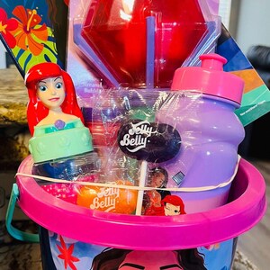 May include: A pink bucket filled with Disney-themed toys and treats. Includes a Moana activity book, a red bubble wand, a purple water bottle, a Jelly Belly lollipop, and an Ariel figurine. The bucket has a pink handle and rim.
