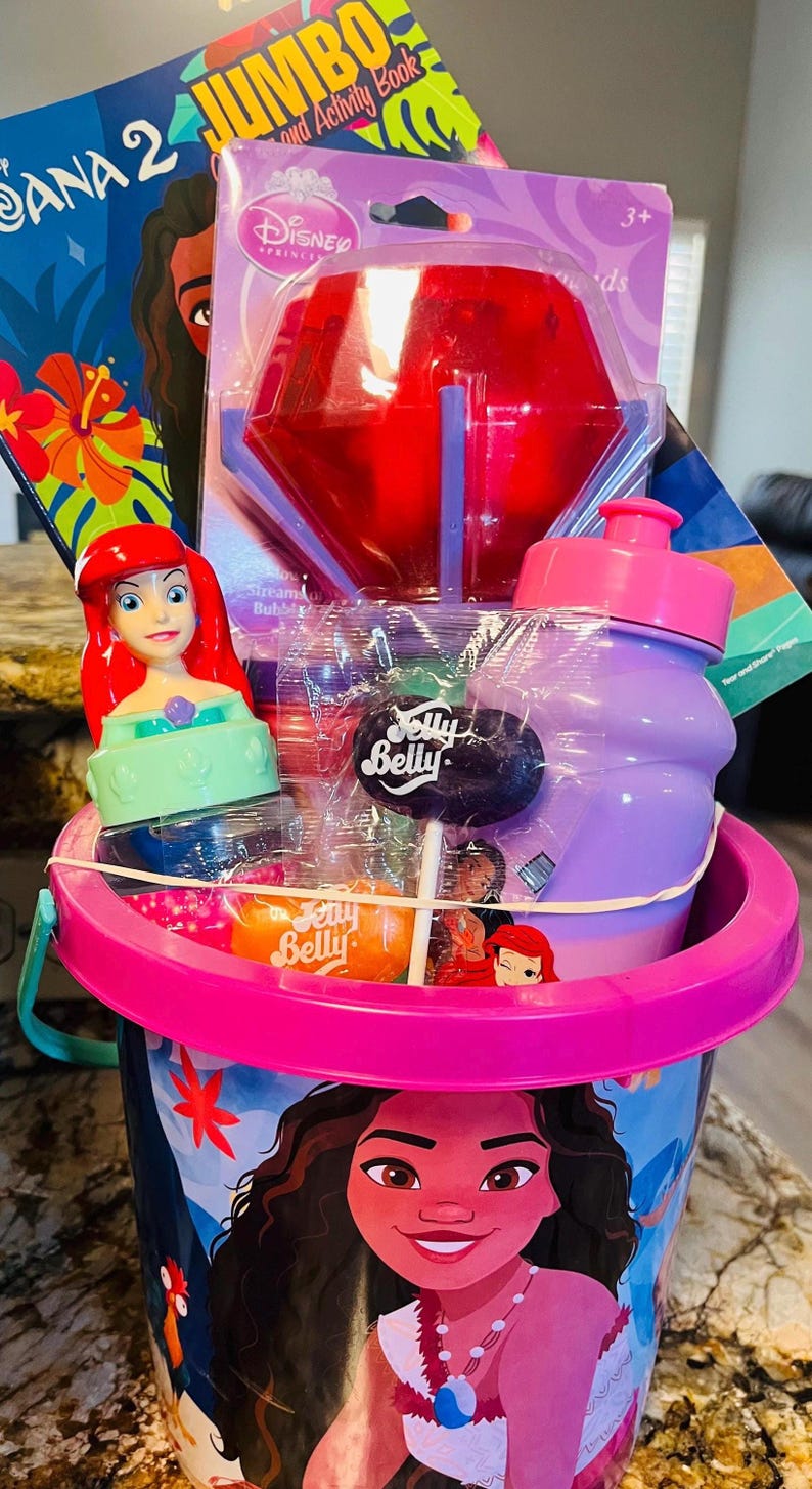May include: A pink bucket filled with Disney-themed toys and treats. Includes a Moana activity book, a red bubble wand, a purple water bottle, a Jelly Belly lollipop, and an Ariel figurine. The bucket has a pink handle and rim.
