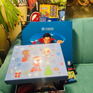 Peut inclure: Une bo&icirc;te bleu clair avec un couvercle orn&eacute; de personnages de Spider-Man en tenues de f&ecirc;te. La bo&icirc;te est ouverte, r&eacute;v&eacute;lant un objet rouge et d'autres articles. Une bo&icirc;te bleue avec une image de Superman est derri&egrave;re, avec un plaid Avengers.