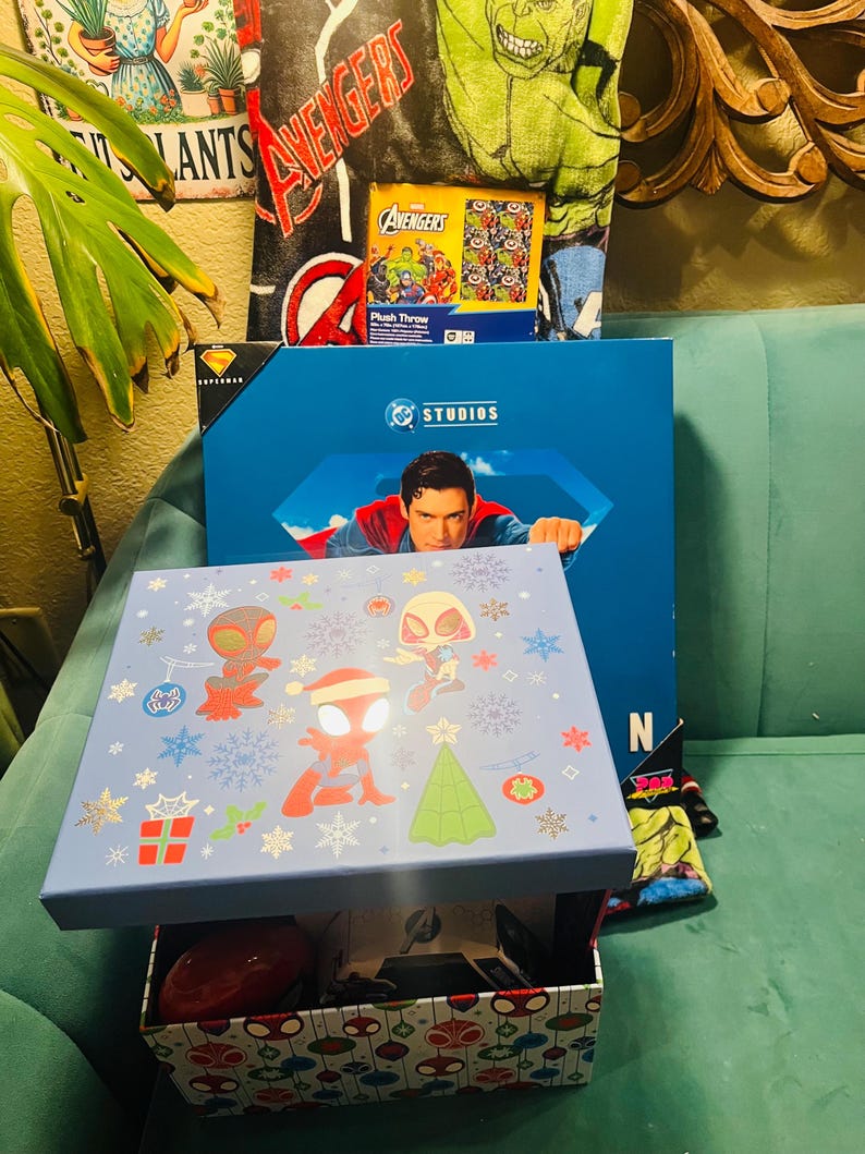 Peut inclure: Une bo&icirc;te bleu clair avec un couvercle orn&eacute; de personnages de Spider-Man en tenues de f&ecirc;te. La bo&icirc;te est ouverte, r&eacute;v&eacute;lant un objet rouge et d'autres articles. Une bo&icirc;te bleue avec une image de Superman est derri&egrave;re, avec un plaid Avengers.