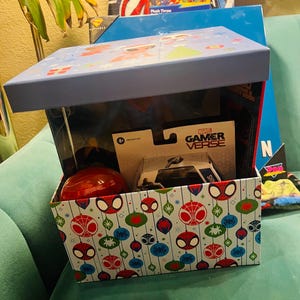 Peut inclure: Une bo&icirc;te cadeau sur le th&egrave;me de Spider-Man, avec un couvercle bleu et un ext&eacute;rieur &agrave; motifs avec des visages et des ornements de Spider-Man. &Agrave; l'int&eacute;rieur se trouvent un jouet rouge, une voiture jouet et d'autres articles.