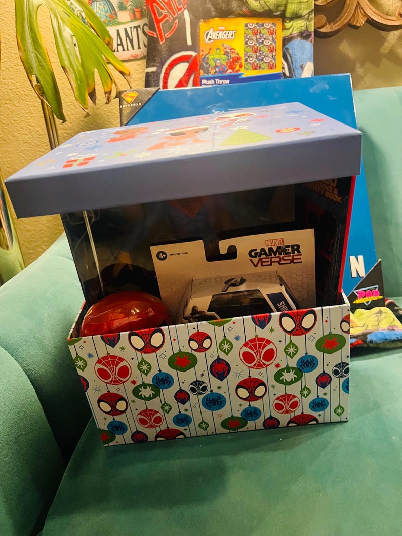 Peut inclure: Une bo&icirc;te cadeau sur le th&egrave;me de Spider-Man, avec un couvercle bleu et un ext&eacute;rieur &agrave; motifs avec des visages et des ornements de Spider-Man. &Agrave; l'int&eacute;rieur se trouvent un jouet rouge, une voiture jouet et d'autres articles.