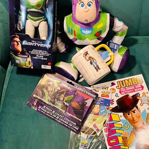 May include: A collection of Disney Pixar Toy Story merchandise, including a Buzz Lightyear action figure in a box, a plush Buzz Lightyear, a Woody mug, a coloring book, crayons, and a paddle ball toy. The items are arranged on a green surface.