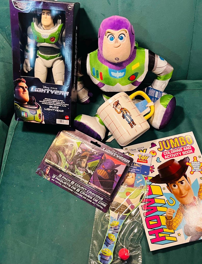 May include: A collection of Disney Pixar Toy Story merchandise, including a Buzz Lightyear action figure in a box, a plush Buzz Lightyear, a Woody mug, a coloring book, crayons, and a paddle ball toy. The items are arranged on a green surface.