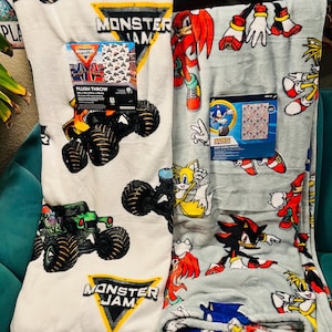 Op de afbeelding: Er worden twee pluche dekens getoond. E&eacute;n is wit met Monster Jam monstertruck graphics en het Monster Jam logo. De andere is lichtgrijs met Sonic the Hedgehog karakters. Beide dekens hebben hun originele verpakking.