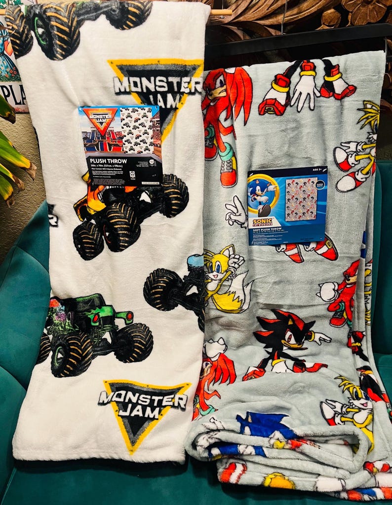 Op de afbeelding: Er worden twee pluche dekens getoond. E&eacute;n is wit met Monster Jam monstertruck graphics en het Monster Jam logo. De andere is lichtgrijs met Sonic the Hedgehog karakters. Beide dekens hebben hun originele verpakking.