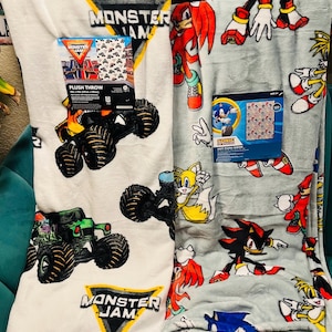 Op de afbeelding: Twee pluche dekens. E&eacute;n is wit met Monster Jam monstertruck graphics. De andere is lichtgrijs met Sonic the Hedgehog-personages. Beide dekens hebben productverpakkingen. De Monster Jam deken heeft een zwart, geel en wit logo.