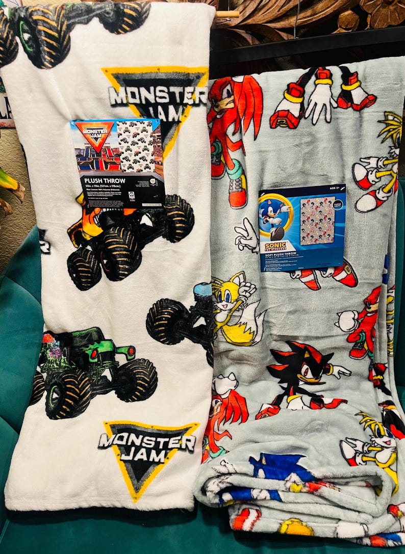 Op de afbeelding: Twee pluche dekens. E&eacute;n is wit met Monster Jam monstertruck graphics. De andere is lichtgrijs met Sonic the Hedgehog-personages. Beide dekens hebben productverpakkingen. De Monster Jam deken heeft een zwart, geel en wit logo.