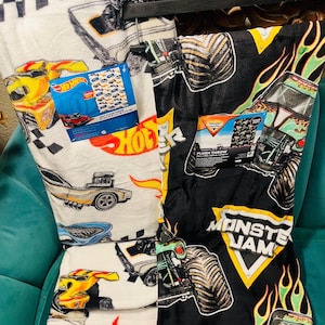 K&ouml;nnte beinhalten: Zwei Pl&uuml;schdecken, eine wei&szlig;e mit Hot Wheels Rennwagen und die andere schwarze mit Monster Jam Monstertrucks. Die Hot Wheels Decke hat ein kariertes Flaggenmuster und die Monster Jam Decke hat Flammen. Beide haben Produktetiketten.