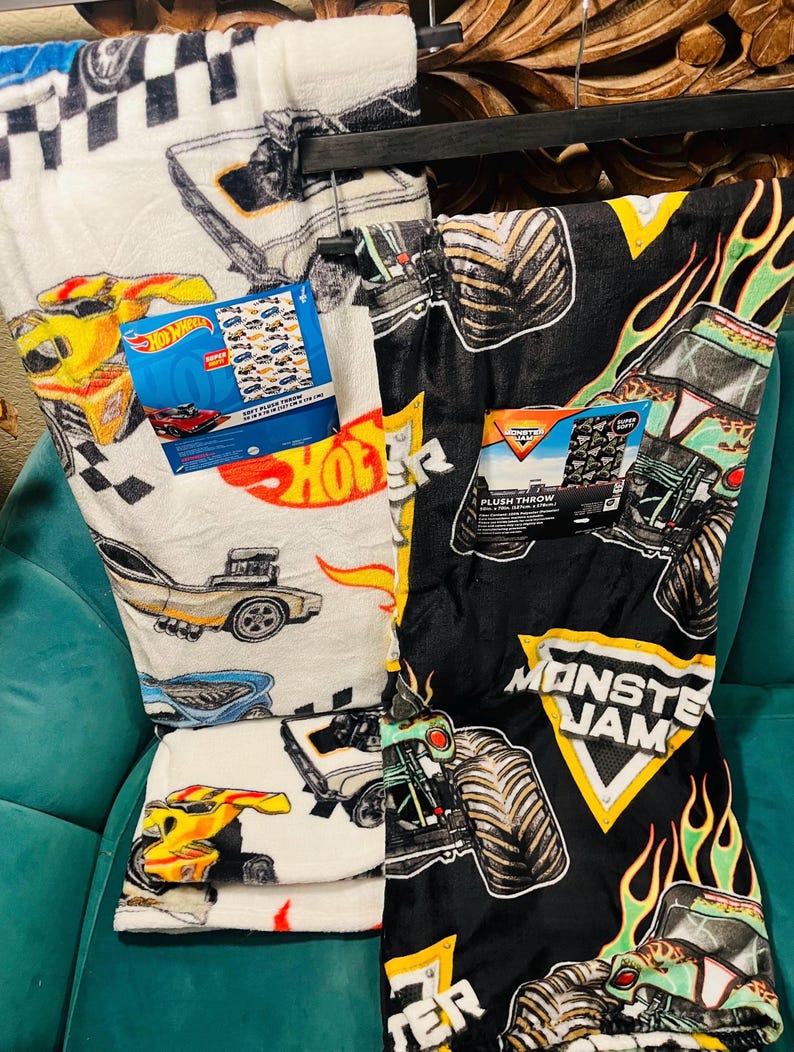 K&ouml;nnte beinhalten: Zwei Pl&uuml;schdecken, eine wei&szlig;e mit Hot Wheels Rennwagen und die andere schwarze mit Monster Jam Monstertrucks. Die Hot Wheels Decke hat ein kariertes Flaggenmuster und die Monster Jam Decke hat Flammen. Beide haben Produktetiketten.