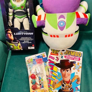 May include: A collection of Toy Story merchandise, including a Buzz Lightyear action figure in a box, a plush Buzz Lightyear, a coloring book, and lollipops. The coloring book features Woody. The items are on a green surface.