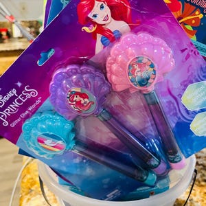 Puede incluir: Un conjunto de varitas m&aacute;gicas Disney Princess Ariel Glitter Dive en un cubo transparente. Las varitas presentan un dise&ntilde;o de concha marina en rosa, morado y azul, con ilustraciones de Ariel. El embalaje incluye el logotipo de Disney Princess y las palabras "Glitter Dive Wands".