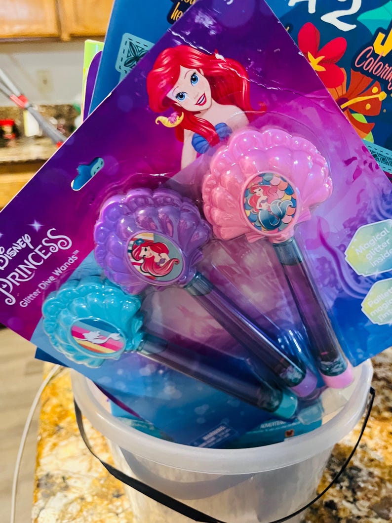 Puede incluir: Un conjunto de varitas m&aacute;gicas Disney Princess Ariel Glitter Dive en un cubo transparente. Las varitas presentan un dise&ntilde;o de concha marina en rosa, morado y azul, con ilustraciones de Ariel. El embalaje incluye el logotipo de Disney Princess y las palabras "Glitter Dive Wands".