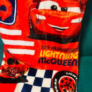 Puede incluir: Manta roja de Lightning McQueen con rayas blancas y un patr&oacute;n de cuadros azules y blancos. La manta presenta una imagen grande de Lightning McQueen y el texto "THE LEGENDARY LIGHTNING McQUEEN".