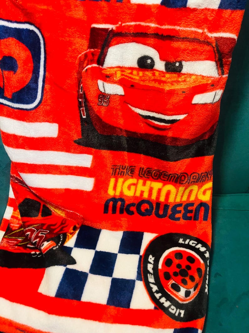 Puede incluir: Manta roja de Lightning McQueen con rayas blancas y un patr&oacute;n de cuadros azules y blancos. La manta presenta una imagen grande de Lightning McQueen y el texto "THE LEGENDARY LIGHTNING McQUEEN".