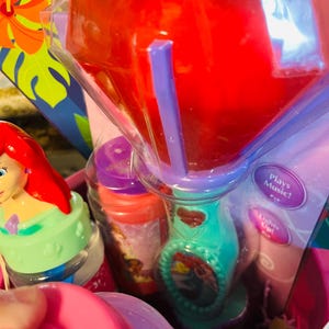 May include: Ariel-themed toy set in a pink basket. Includes a teal and purple toy with a red liquid inside, a small bottle, and a toy figure. The toy has the text "Plays Music!" and "Lights Up!"