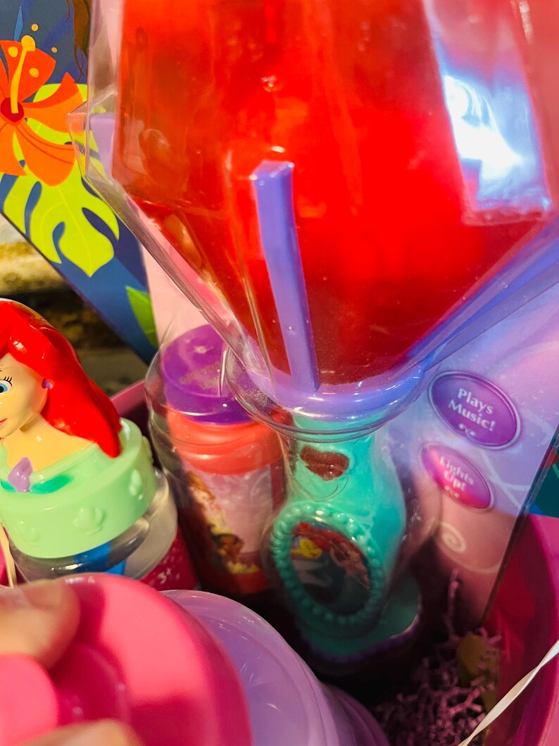 May include: Ariel-themed toy set in a pink basket. Includes a teal and purple toy with a red liquid inside, a small bottle, and a toy figure. The toy has the text "Plays Music!" and "Lights Up!"