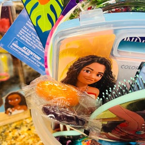 Puede incluir: Un colorido set de regalo con un dise&ntilde;o del personaje Moana. El set incluye un recipiente azul claro, un cepillo para el pelo y otros art&iacute;culos. La imagen tambi&eacute;n muestra un peque&ntilde;o objeto naranja envuelto y una peque&ntilde;a figura de Moana. El texto "ATTENTION!" es visible.