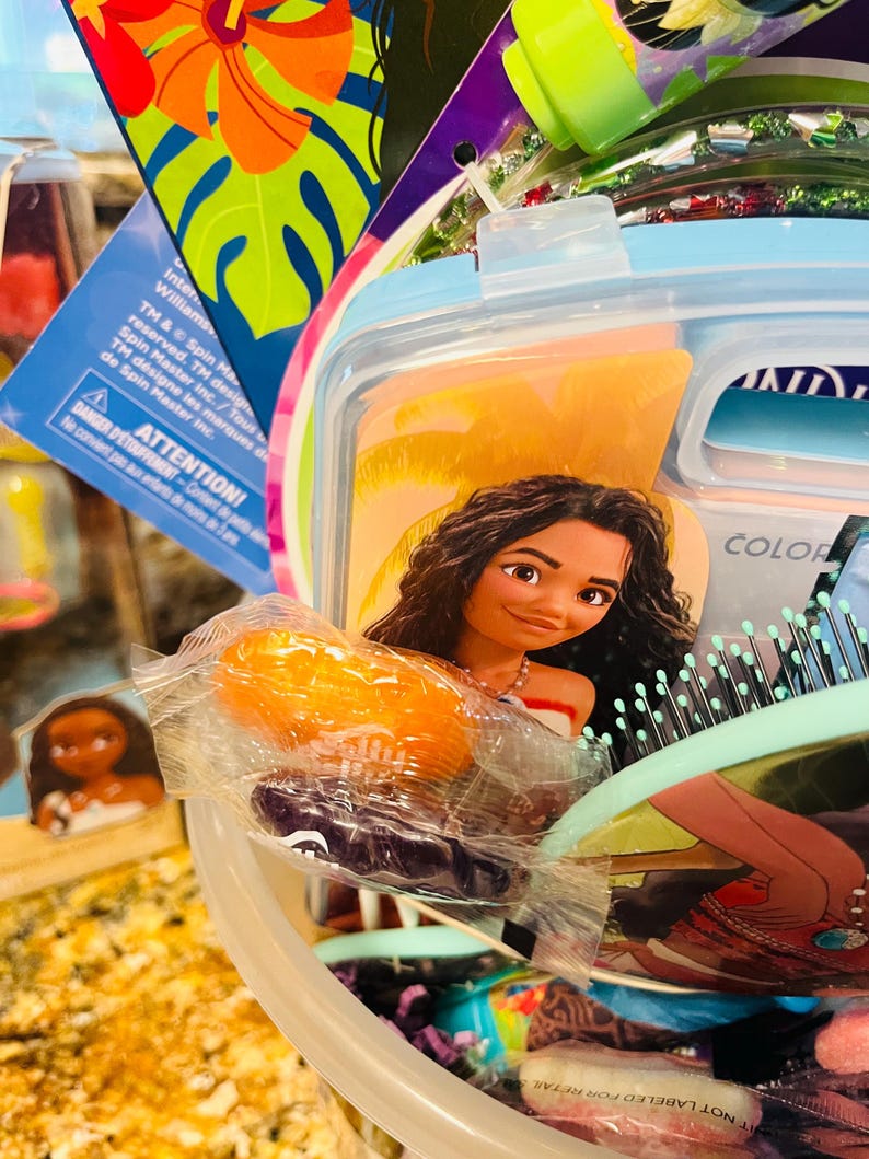 Puede incluir: Un colorido set de regalo con un dise&ntilde;o del personaje Moana. El set incluye un recipiente azul claro, un cepillo para el pelo y otros art&iacute;culos. La imagen tambi&eacute;n muestra un peque&ntilde;o objeto naranja envuelto y una peque&ntilde;a figura de Moana. El texto "ATTENTION!" es visible.