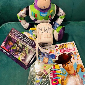 May include: A collection of Toy Story themed items, including a Buzz Lightyear plush, a mug with "HOWDY" printed on it, a coloring book, a paddle ball, and a box of crayons. The items are arranged on a green surface.