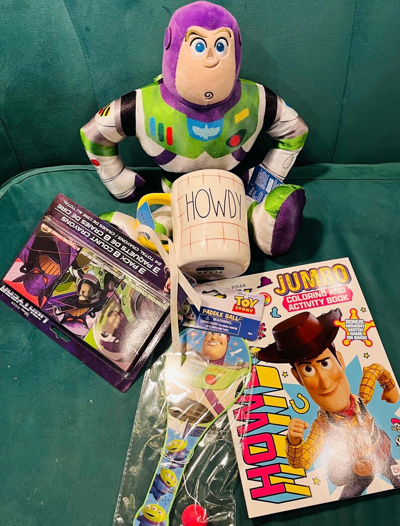 May include: A collection of Toy Story themed items, including a Buzz Lightyear plush, a mug with "HOWDY" printed on it, a coloring book, a paddle ball, and a box of crayons. The items are arranged on a green surface.