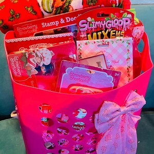 May include: A pink basket filled with Strawberry Shortcake themed items. The basket contains a plush throw, toys, and a bow. The items include a stamp set, slime, and a face mask. The basket is on a blue surface.