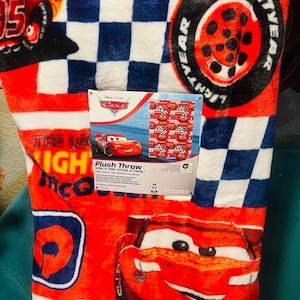 Puede incluir: Manta de felpa roja con Lightning McQueen de Cars de Disney Pixar. La manta tiene un patr&oacute;n de cuadros en azul y blanco, y el texto "LIGHTYEAR" es visible. La manta mide 127 cm por 178 cm.