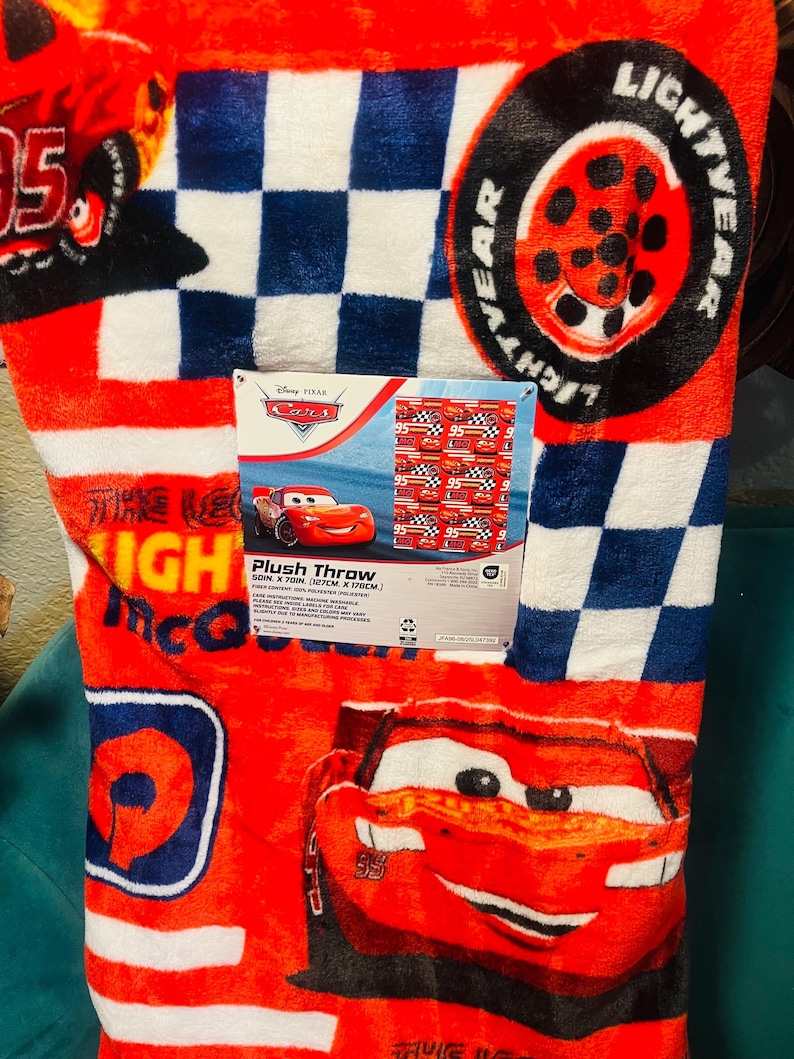 Puede incluir: Manta de felpa roja con Lightning McQueen de Cars de Disney Pixar. La manta tiene un patr&oacute;n de cuadros en azul y blanco, y el texto "LIGHTYEAR" es visible. La manta mide 127 cm por 178 cm.