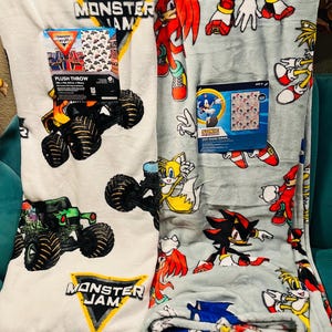Op de afbeelding: Er worden twee pluche plaids getoond. E&eacute;n is wit met monster truck-afbeeldingen en de tekst "Monster Jam". De andere is lichtgrijs met Sonic the Hedgehog-personages. Beide plaids hebben een aangehechte productverpakking.