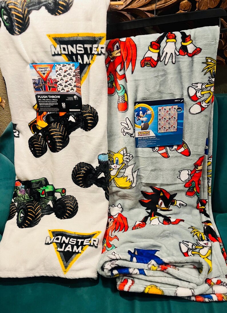 Op de afbeelding: Er worden twee pluche plaids getoond. E&eacute;n is wit met monster truck-afbeeldingen en de tekst "Monster Jam". De andere is lichtgrijs met Sonic the Hedgehog-personages. Beide plaids hebben een aangehechte productverpakking.