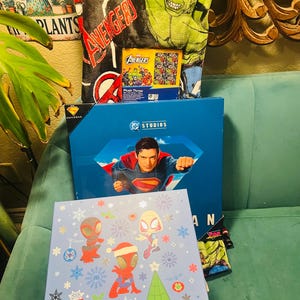 Peut inclure: Ensemble d'articles sur le th&egrave;me des super-h&eacute;ros, comprenant une couverture Avengers, une bo&icirc;te Superman et une bo&icirc;te bleu clair avec des personnages de Spider-Man et des d&eacute;corations de No&euml;l. Le tout est dispos&eacute; sur un canap&eacute; turquoise.