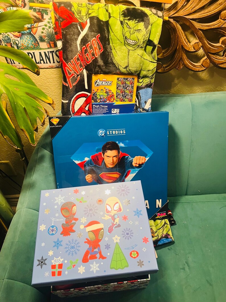 Peut inclure: Ensemble d'articles sur le th&egrave;me des super-h&eacute;ros, comprenant une couverture Avengers, une bo&icirc;te Superman et une bo&icirc;te bleu clair avec des personnages de Spider-Man et des d&eacute;corations de No&euml;l. Le tout est dispos&eacute; sur un canap&eacute; turquoise.