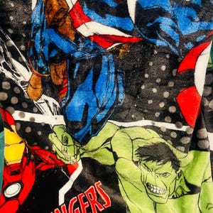 Peut inclure: Une couverture en peluche avec un design vibrant des personnages des Avengers. La couverture pr&eacute;sente Captain America, Iron Man, Hulk et d'autres h&eacute;ros dans des poses d'action sur un fond noir avec le mot "Avengers" en rouge.