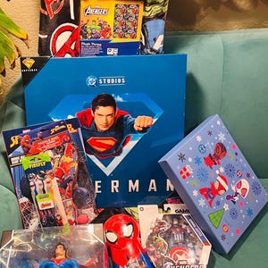 Peut inclure: Une collection de produits d&eacute;riv&eacute;s sur le th&egrave;me des super-h&eacute;ros, comprenant une couverture en peluche Avengers, un coffret Superman, des jouets Spider-Man et une figurine Avengers Captain America. Les articles sont dans diverses bo&icirc;tes et emballages, avec des couleurs vives et des motifs de personnages.