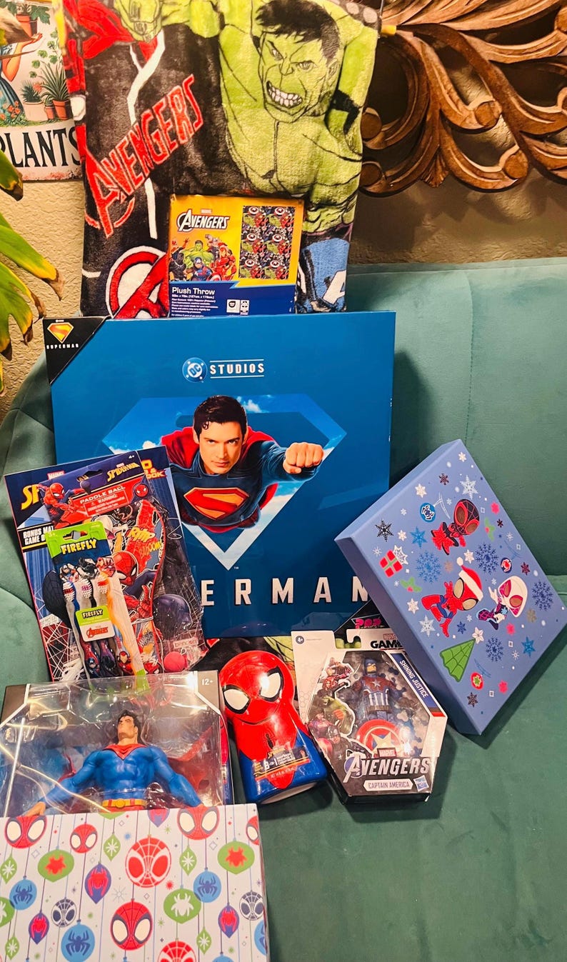 Peut inclure: Une collection de produits d&eacute;riv&eacute;s sur le th&egrave;me des super-h&eacute;ros, comprenant une couverture en peluche Avengers, un coffret Superman, des jouets Spider-Man et une figurine Avengers Captain America. Les articles sont dans diverses bo&icirc;tes et emballages, avec des couleurs vives et des motifs de personnages.