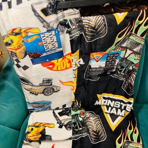 K&ouml;nnte beinhalten: Zwei Pl&uuml;schdecken, eine wei&szlig;e mit Hot Wheels-Autografiken und die andere schwarze mit Monster Jam-Monstertruck-Grafiken. Beide Decken haben angebrachte Etiketten. Die Hot Wheels-Decke zeigt eine Vielzahl von Rennwagen.