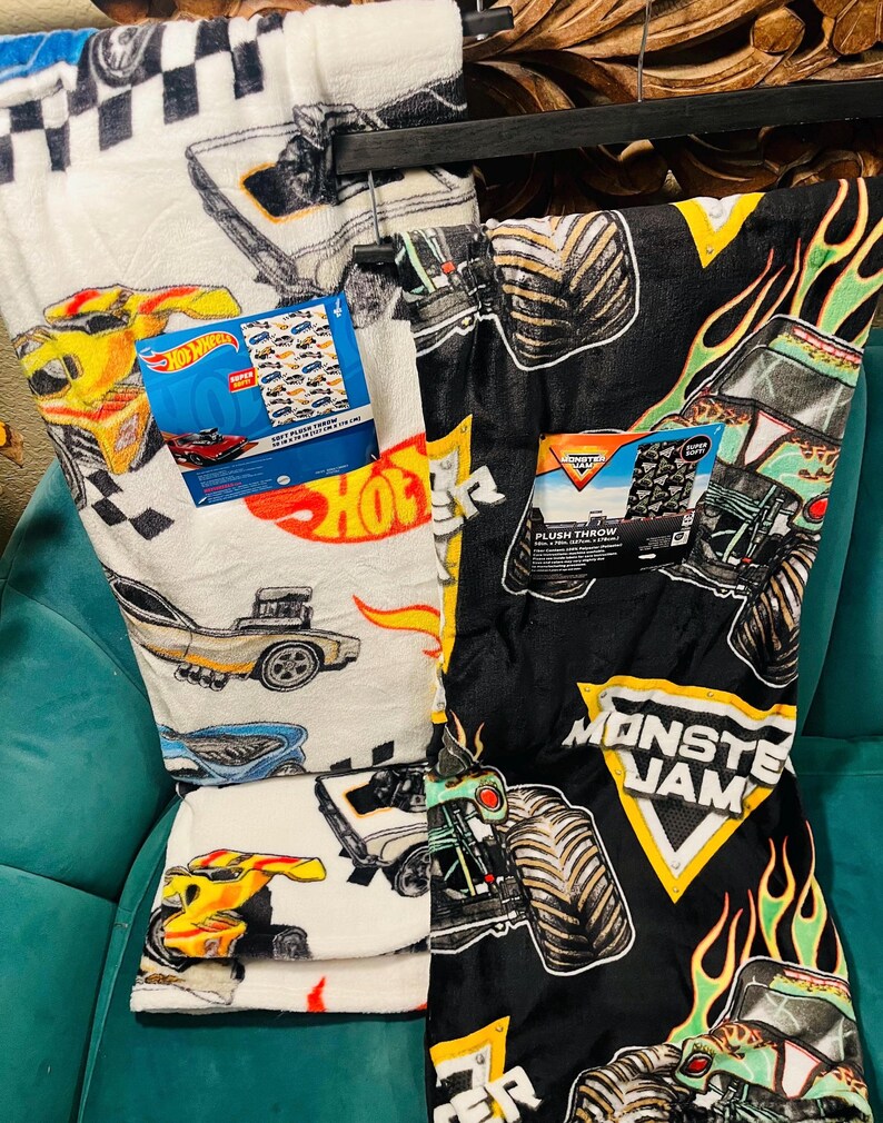 K&ouml;nnte beinhalten: Zwei Pl&uuml;schdecken, eine wei&szlig;e mit Hot Wheels-Autografiken und die andere schwarze mit Monster Jam-Monstertruck-Grafiken. Beide Decken haben angebrachte Etiketten. Die Hot Wheels-Decke zeigt eine Vielzahl von Rennwagen.