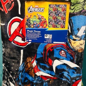 Peut inclure: Couverture en peluche avec les personnages des Avengers de Marvel, dont Hulk et Captain America, dans un style bande dessin&eacute;e. La couverture est noire avec des graphismes rouges, bleus, verts et blancs. Une bo&icirc;te avec le logo des Avengers est visible.