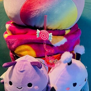 May include: A plush toy arrangement featuring a large lavender cat-shaped plush with a rainbow belly, a pink blanket, and two smaller plush toys in a decorative cup. A pink candy-shaped straw adds a playful touch.