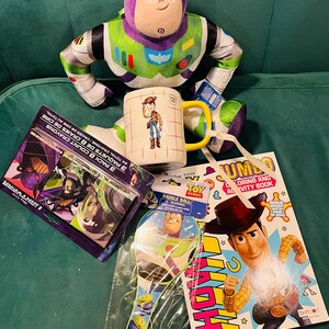 May include: A collection of Toy Story themed items, including a Buzz Lightyear plush, a Woody mug, a coloring book, a paddle ball, and crayon packs. The items are arranged on a green surface.