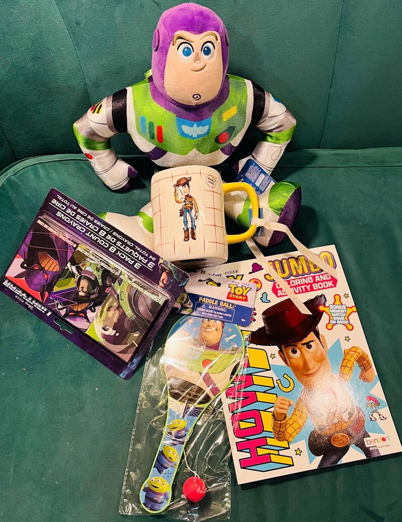 May include: A collection of Toy Story themed items, including a Buzz Lightyear plush, a Woody mug, a coloring book, a paddle ball, and crayon packs. The items are arranged on a green surface.