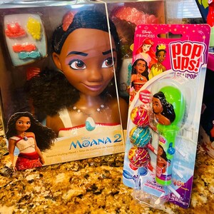 May include: A Moana styling head doll and a package of lollipops featuring Moana and other Disney princesses. The doll is in a box with hair accessories. The lollipops are in a clear plastic package with a green handle.