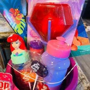May include: A pink Easter basket filled with Disney Princess-themed items. Includes a red light-up toy, a water bottle, lollipops, and a bottle with Ariel on it. A Moana book is visible in the background.