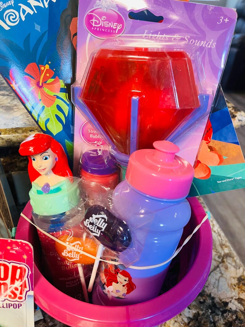 May include: A pink Easter basket filled with Disney Princess-themed items. Includes a red light-up toy, a water bottle, lollipops, and a bottle with Ariel on it. A Moana book is visible in the background.