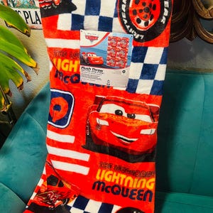 Puede incluir: Manta de felpa roja con Lightning McQueen de la pel&iacute;cula Cars. La manta tiene un patr&oacute;n de cuadros en blanco y azul marino, con gr&aacute;ficos de Lightning McQueen y neum&aacute;ticos. Tambi&eacute;n es visible el texto "The Legendary Lightning McQueen".